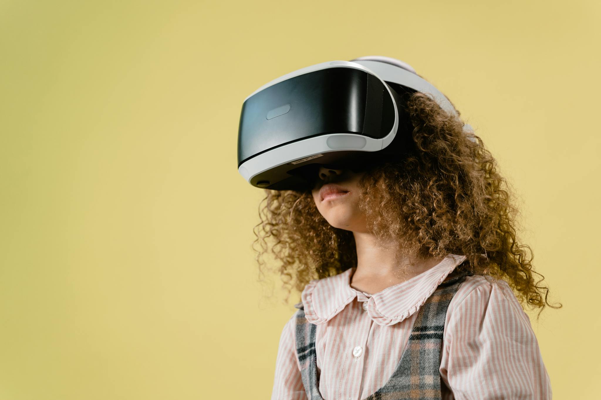 A child with curly hair wearing VR goggles against a yellow background. Ideal for tech and lifestyle themes.